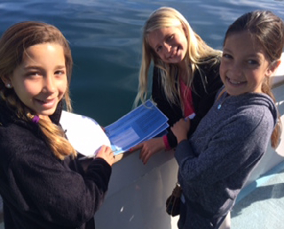 Three young girls stand on a boat on the Pacific Ocean and look through a booklet during the Marine Education program.